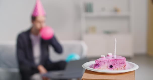 Close-up of slice of pink birthday cake with candle lies on white plate stands in living room against home interior background in blurred unfocused human silhouette man sits on couch inflates balloon — Stock Video