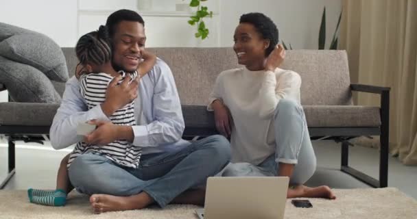 Afroamerikansk ung familie sidder hjemme på gulvet på hvidt tæppe med bærbar computer i weekenden, kærlig far krammer sin lille bedårende datter krammer søde barn, kærlige forældre med barn slapper af i nyt hjem. – Stock-video