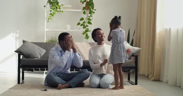 Familie spielt Verstecken auf dem Boden im gemütlichen Wohnzimmer, afroamerikanische Eltern verbringen am Wochenende Zeit mit ihrer Tochter, bedecken ihre Augen mit den Händen und tun so, als ob sie unsichtbar wären — Stockvideo