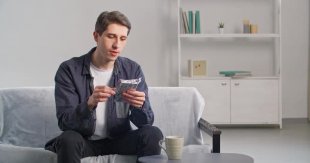 En ung mann sitter på sofaen hjemme med en innpakket sjokoladeplate i hendene og åpner dessertsnus aromatisk duft av kakaobitt uttrykker glede av smak nyter sødme under frokost eller lunsj – stockvideo