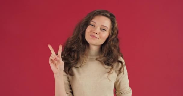 Kobieta studio portret kaukaski millenium dziewczyna patrząc na aparat pozowanie na czerwonym tle w casual ubrania pokazując gest pokoju drugi numer z dwoma palcami symbol przyjaźni znak zwycięstwa — Wideo stockowe