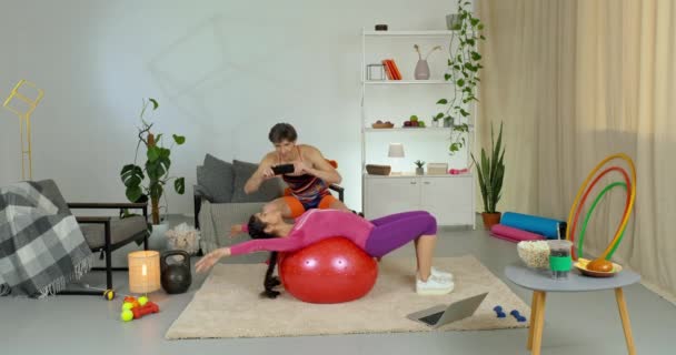 Slender εκπαιδευτής κορίτσι βρίσκεται στην πλάτη στο κόκκινο μπάλα σπορ κάνει ασκήσεις κατάρτισης με fitball στο σπίτι τέντωμα πτώση στο πάτωμα, ενώ ο άνθρωπος παίρνει φωτογραφία βίντεο γυρίσματα προπόνηση στο τηλέφωνο streaming στο blog — Αρχείο Βίντεο