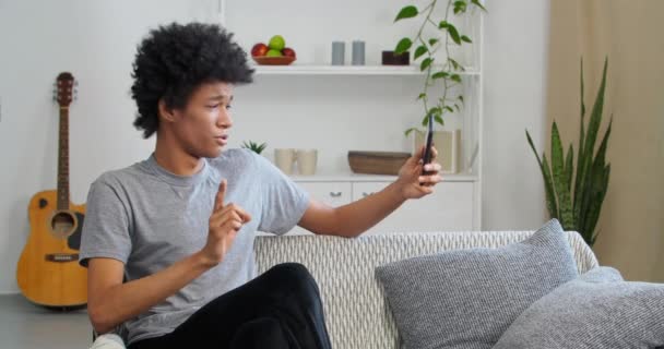 Afro Američan chlap teenager sedí na gauči doma v obývacím pokoji drží telefon v ruce mluví do smartphone kamery dělá on-line záznam hovoru video pro blog odmítá otázku vlny prst nesouhlasně — Stock video