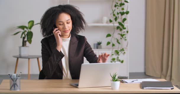 Afro amerikanska affärskvinna lockigt flicka frilansare arbetar med bärbar dator på distans på kontoret svar samtal talar på mobiltelefon med vän konsulter leenden njuter av samtal med wirelles enhet — Stockvideo