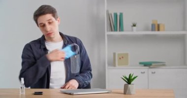 Masasında oturan beyaz adam tıbbi koruyucu maskesini çıkarıyor virüs mikroplarından elde antiseptik alıyor ev bitkisine ve kafasına alkol püskürtüyor ve ağzını dezenfekte ediyor.