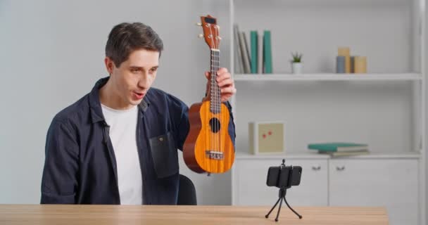 Homem confiante blogueiro grava vídeo vlog em casa sentado à mesa usando celular mostra ukulele tocando guitarra pequena instrumento musical melodia musical faz performance online durante quarentena — Vídeo de Stock