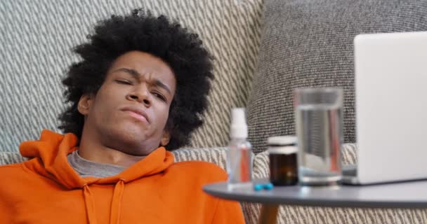 Portrait of curly-haired guy teenager wears orange sweater suffers from discomfort in throat and body feels pain coughs at home watching movies on laptop online during epidemic of respiratory virus — Stock Video