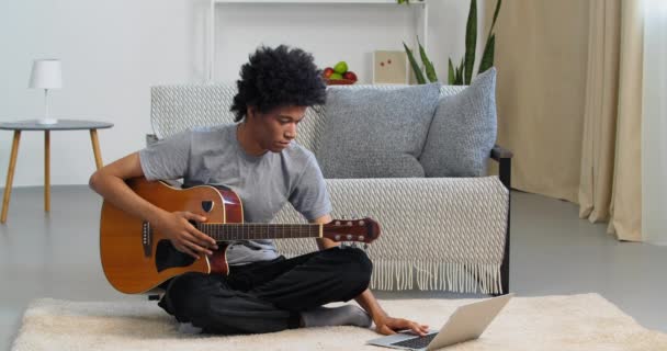 Этнический подросток смотрит онлайн-видеоурок музыки на ноутбуке, учится играть на гитаре дома, африканский молодой человек практикует игру на музыкальных инструментах с помощью компьютерного приложения для интернет-учителей — стоковое видео
