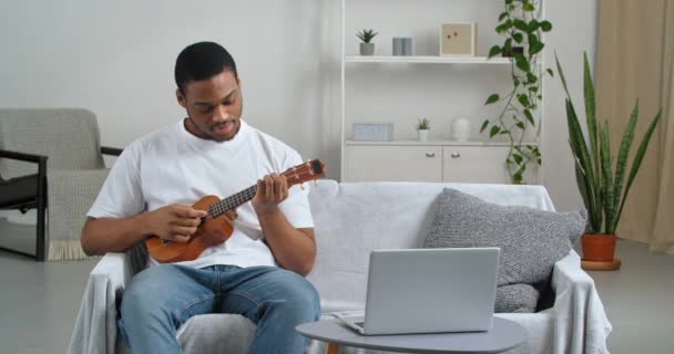 Афро-американец носит белую футболку и джинсы, сидя в уютной гостиной на диване, наблюдая уроки музыкального видео онлайн, используя ноутбук, учась играть на маленьком укулеле, радуется, делая победный жест рукой — стоковое видео