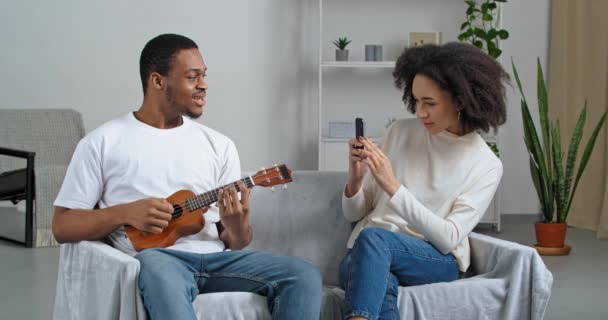 Afro amerykański mężczyzna muzyk siedzi na kanapie śpiewając małe hawajskie gitara instrument muzyczny dziewczyna kręcone kobieta za pomocą telefonu komórkowego pomaga chłopak nagrywać wideo muzyka lekcja robienia zdjęć — Wideo stockowe