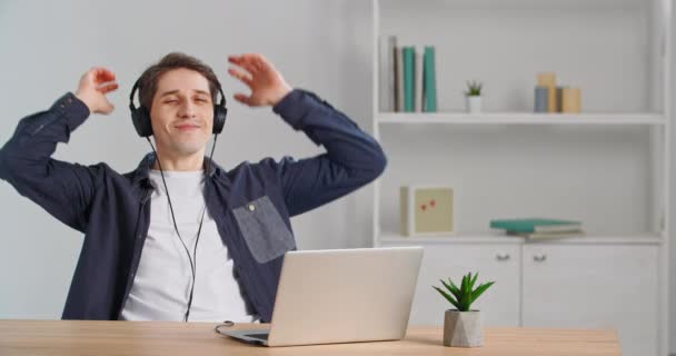 Happy funny caucasian man sitting at table in office listening to music online wearing wireless headphones enjoying favorite track song emotionally pretending playing guitar having fun at workplace — Stock Video