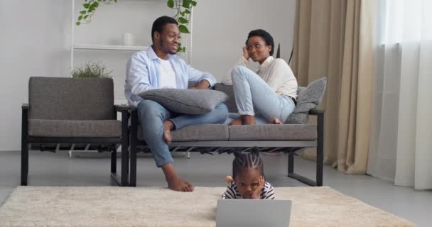 Jóvenes padres responsables afroamericanos pareja americana sentada en el sofá hablando mientras que la niña preescolar mira la pantalla del ordenador portátil navegando por la red jugando juegos de ordenador que estudian de forma remota — Vídeo de stock