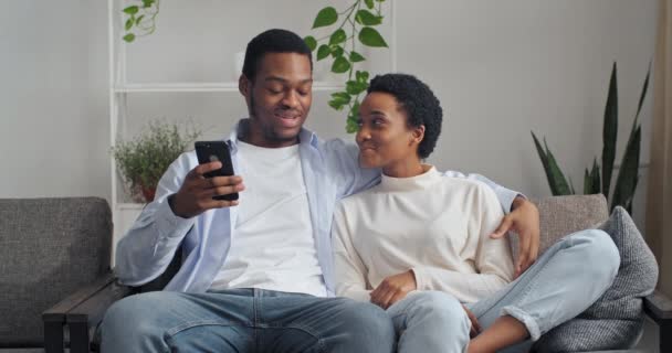 Amar a la pareja étnica afroamericana sentada en el sofá en casa hablando de pasar tiempo juntos en las vacaciones. Carrera mixta sonrió hombre y mujer relajándose en el sofá con la comunicación de teléfonos inteligentes en la red social — Vídeos de Stock