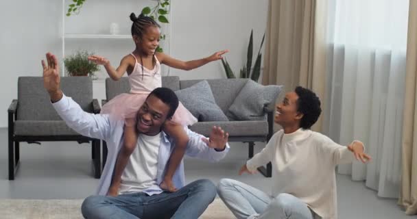 Αφρο-αμερικανοί γονείς με την κόρη τους να κάθεται στο πάτωμα στο σαλόνι ο πατέρας κρατώντας το κοριτσάκι στο πίσω μέρος του λαιμού του χορεύοντας μαζί εθνοτική οικογένεια κάνοντας κινήσεις κυμάτων με τα χέρια προσποιούνται αεροπλάνο — Αρχείο Βίντεο