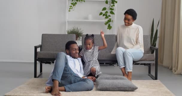Afro amerikansk familie tilbringer tid i hyggelig stue, lille sjov pige fortæller hemmelighed i hendes forældre øre for at dele fortrolige oplysninger sladder spiller med mor og far griner aktivt fagter samtaler – Stock-video