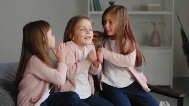 Three little girls sisters in same clothes sitting on couch at home telling secrets to each other, two children kids funny whispering confidential private information gossip in ear of female friend — Stock Video