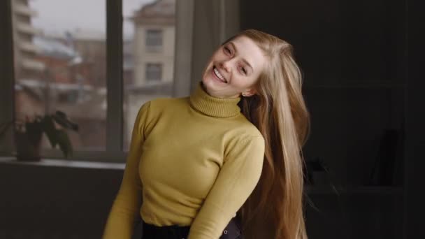 Jong Kaukasisch meisje vrouw millennial vrouw model draagt geel stijlvolle trui staat thuis poseren geïsoleerde gooit lang recht blond haar in de lucht speelt met kapsel lacht kijken naar camera — Stockvideo