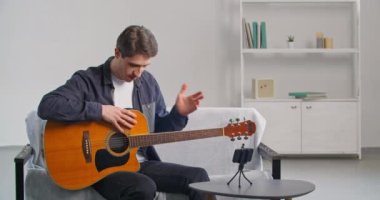 Gündelik kıyafetli genç öğretmen evde kanepede oturmuş müzik enstrümanının yapısını anlatan gitar çalma üzerine online ders Webinar video dersi veriyor.