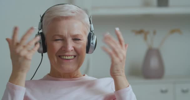 Elderly woman caucasian grandmother pensioner lady puts on headphones modern device enjoys fly listens to songs sincerely smiles dental moves her hands dancing feels happiness delight sitting at home — Stock Video