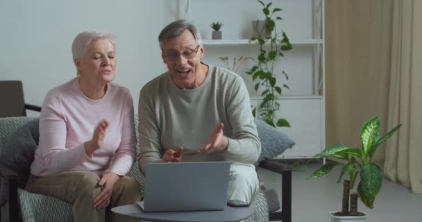Бабусі і дідусі сидять на дивані вдома в інтер'єрі, розмовляючи в веб-камеру сучасного ноутбука, махаючи руками вітання, кажучи привіт і прощання, виступаючи на відеоконференції онлайн дзвінки з онуками — стокове відео