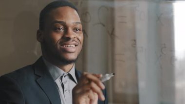 Yalnız Afro-Amerikalı iş adamı erkek lider modern ofiste duruyor, fikirler formülü yazıyor, cam şeffaf tahtada finans kârı sayıyor, stratejileri analiz ediyor, iyi sonuçlarla gülümsüyor.
