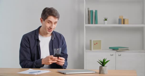 Feliz hombre adulto joven usuario joven sentado en la mesa sosteniendo el teléfono inteligente en sus manos, recibir un mensaje en la red social, respuestas escribiendo, sonriendo pensativo, utiliza la aplicación móvil para la comunicación de chat en el teléfono — Vídeos de Stock