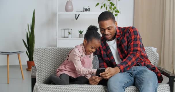 愛する父は小さな娘の就学前アフロアメリカの女の子とソファの上に座っているスマートフォンで、単一の親の家族は、インターネット上で一緒に楽しみを持っている漫画は、モバイルデバイスでオンラインゲームをプレイ — ストック動画