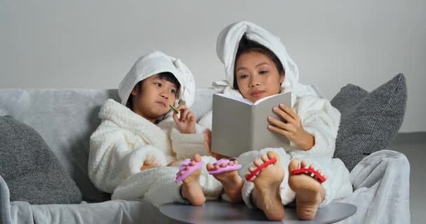 Молода азіатська мати розслабляється на дивані з маленькою донькою їсть печиво солодощі дівчата використовують розширювач пальців для педикюру час з процедурою ніг жінка читає сіру книжкову літературу вдома на дивані — стокове відео