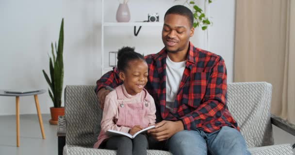 Afro americano padre etnico insegnante maggiore fratello insegna poco divertente ragazza a leggere bambino in possesso di grigio libro mostra figlia foto album ridere parlando parenti trascorrere del tempo seduti sul divano, educazione a casa — Video Stock