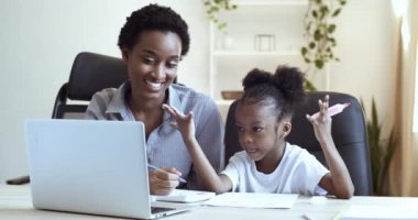 Genç anne Afro-Amerikan bayan öğretmen ve tatlı kızı okullu siyah çocuk dizüstü bilgisayarda oturuyor uzaktan, uzaktan, internette ödev yapıyor ve yakından gülüyor.