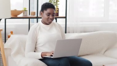 Afro-Amerikalı serbest çalışan kadın iş kadını, evdeki koltukta oturuyor, online alışveriş için bilgisayar uygulaması kullanıyor, uzaktan çalışıyor. Etnik siyah Afrikalı milenyum kızı laptopunu içeride yazıyor.