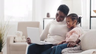 Afro-Amerikan anne anne kadın ve küçük zenci Afrikalı kız çocuk dizüstü bilgisayar uygulaması kullanarak evde oturuyor, webcam ile konuşuyor, online sohbet konferansı yapıyor, çevrimiçi video izliyorlar.