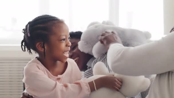 Afro amerikansk familj spelar spel med leksaksbjörn hemma soffan skrattar tillsammans. Ung mor svart kvinna äldre syster med liten dotter flicka barn och oigenkännlig man far har kul inomhus — Stockvideo