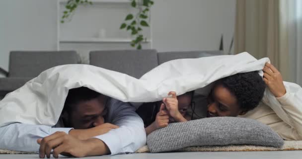 Famille afro-américaine, père africain adulte, jeune mère ethnique, petite fille fille noire enfant couché sur le canapé à la maison se cachant sous une couverture blanche en regardant la caméra sourire montrant comme un pouce vers le haut — Video