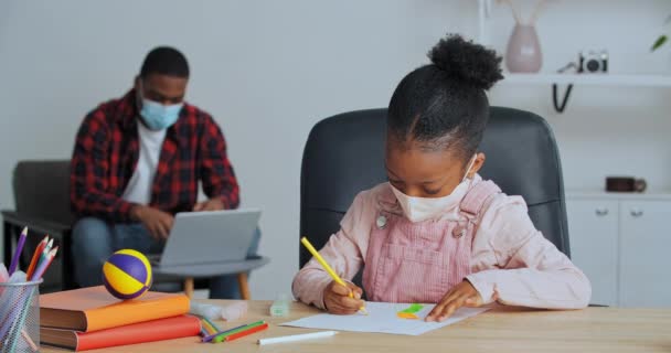 Отец и дочь носят медицинские маски для лица дома, девочка рисует в то время как папа работает на ноутбуке, афро-американец учитель серфинга сети, ребенок делает домашнее задание указывает пальцем к мячу на стол говорить, блокировка — стоковое видео