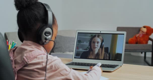 Vista desde atrás afro-americana niña niña lleva auriculares mira en el ordenador portátil en la mujer profesora habla se comunica en línea con la maestra dibuja relojes de vídeo en la red sentado en casa — Vídeo de stock