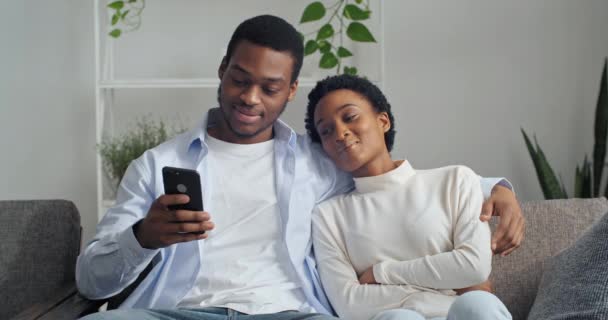 Retrato de duas pessoas afro-americano casal namorado e namorada homem e mulher sorrindo abraço sentado juntos no sofá olhando para o telefone falando ler notícias na rede assistindo vídeo online — Vídeo de Stock