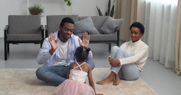 Afro amerykański czarny etniczny rodzina siedzi w domu na podłodze w salonie młodzi rodzice mama i tata z córką w domu zabawy, mała dziewczynka dziecko w różowej sukience oszukiwanie tata o dać pięć rąk — Wideo stockowe