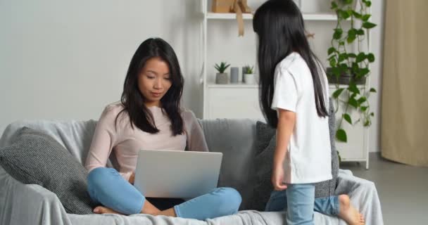 Asiática joven madre soltera freelancer trabaja remotamente desde casa se sienta en el sofá con el ordenador portátil de compras en línea, pequeña niña colegiala hace ruido sacude mamá atraer la atención distrae de los negocios — Vídeo de stock