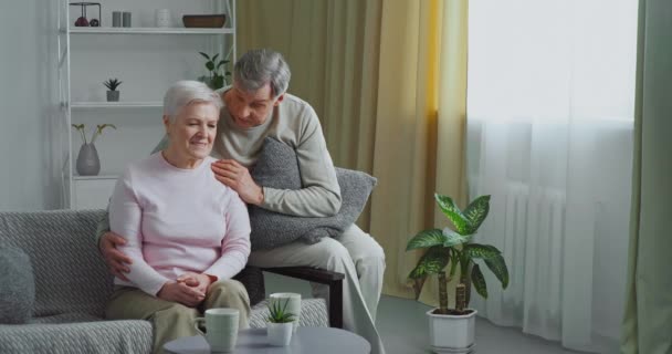 Elderly caucasian couple sits on gray sofa, old man loving husband hugs his wife comforts beloved sad woman feels sorry apologizes supports partner in difficult situation gives psychological help — Stock Video