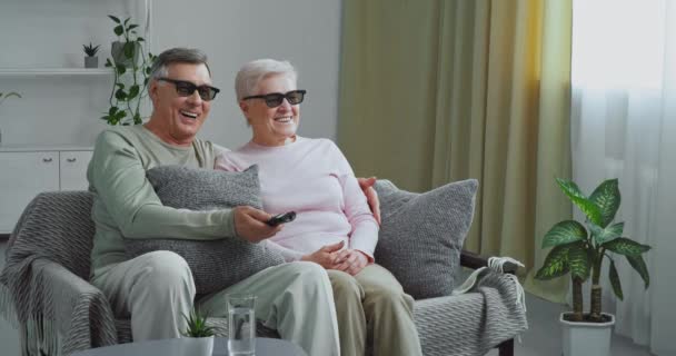 Casal de idosos avós de cabelos grisalhos abraçando sentado no sofá na sala de estar moderna usando óculos 3D tridimensionais assistindo filme engraçado rindo desfrutando de tecnologias modernas de cinema em casa — Vídeo de Stock