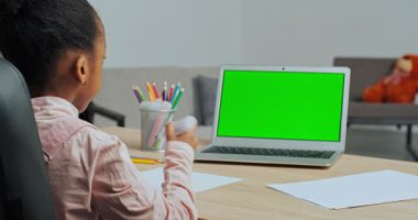 Afro-Amerikan liseli küçük kız masasında oturmuş renkli kalemlerle resim çiziyor yeşil ekran dizüstü bilgisayara, online derse, karantinada uzaktan öğrenmeye bakıyor.