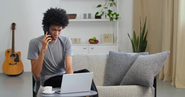 Sérieux afro-américain homme d'affaires étudiant gars parlant sur téléphone portable utilisation ordinateur portable pour le travail à distance étudier à partir du bureau à la maison. Freelancer consulte par smartphone communique en ligne à l'intérieur de l'appartement — Video