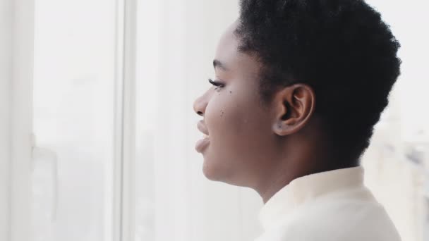 Jovem afro-americana a olhar pela janela. Focada senhora étnica negra africana desfrutando de bela vista e sonhando em casa. Millennial menina contemplando sentindo esperança pacífica em pé no quarto — Vídeo de Stock