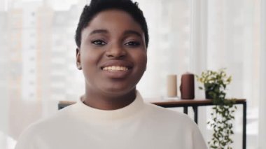 Afro-Amerikan milenyum kızı güzel, 20 'li yaşlarda, etnik kadın mutlu, ırk melezi, kısa saçlı, siyah afro kadın kameraya bakıp evde beyaz odada gülümseyerek poz veriyor.