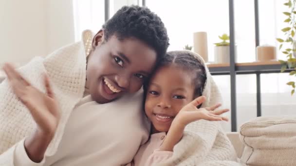 Afro amerikansk mamma mor afrikan kvinna vuxen ung förälder kramar kramar älskad liten flicka svart blandras dotter barn viftande händer hej tillsammans tittar på kameran täckt med filt hemma — Stockvideo
