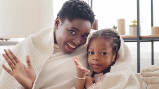 Afro-americana mãe mãe irmã mais velha babá e criança menina filha criança criança negra criança africana estudante sentado em casa coberto com cobertor acenando mãos Olá sorrindo olhando para a câmera — Vídeo de Stock