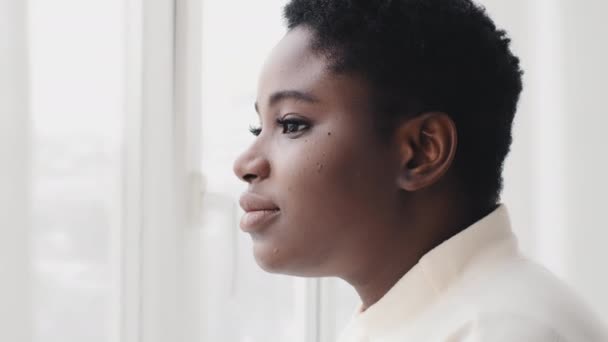 Portret afro amerykańskiej kobiety etnicznej pani millenium czarny dziewczyna afrykański model patrząc przez okno stojąc w świetle dziennym śniąc myśląc kontemplując zamienia twarz kobiety patrząc na aparat uśmiechnięty — Wideo stockowe