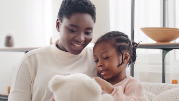 Afro amerikansk mor med liten dotter flicka sitter på soffan hemma pratar håller nalle. Ensamstående etnisk svart mamma blandad ras äldre syster kvinna och barn kvinna barn chatta inomhus — Stockvideo