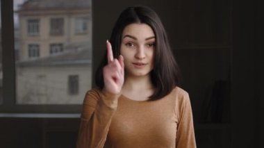 20 'li yaşlardaki genç kız modelinin portresi. Mutlu kadın. Kapalı mekanda durup kamera önünde işaret parmağına bakıyor.